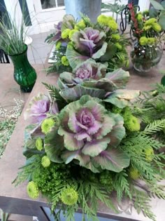 some green and purple flowers on a table with other plants in vases behind them