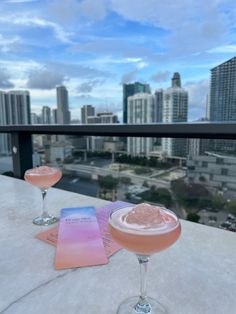 two cocktails sitting on top of a white table next to each other in front of a cityscape