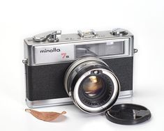 an old fashioned camera sitting next to a small leaf on a white surface with the lens pointed up