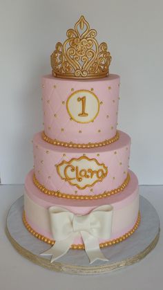 a pink and gold cake with a white bow on the bottom tier is decorated with an ornate crown