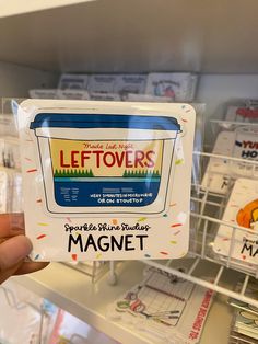 someone holding up a magnet that says leftovers in front of a store shelf filled with magnets