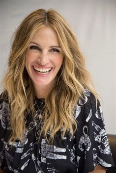 a smiling woman with long blonde hair sitting in front of a white wall and wearing a black shirt