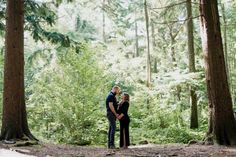 a man and woman standing in the middle of a forest with tall trees behind them