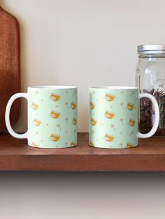 two coffee mugs sitting next to each other on a wooden shelf in front of a glass jar