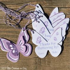 two purple and white butterfly shaped tags sitting on top of a wooden table next to string lights