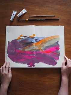 two hands holding an open book on top of a table with paint and crayons