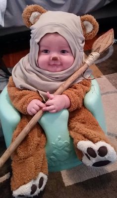 a baby in a bear costume is sitting on a chair with a broom and teddy bear