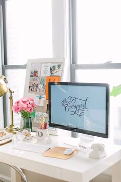 a desktop computer sitting on top of a white desk next to a lamp and flowers