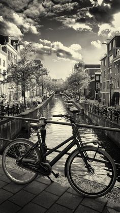 black and white photograph of a bicycle parked on the side of a canal with buildings in the background