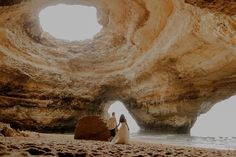 two people sitting on the beach in front of a cave