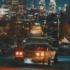 a car driving down the road in front of a city skyline at night with lights on