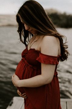 a pregnant woman in a red dress standing on a dock next to the water looking down at her belly