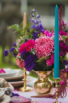 a gold vase filled with purple and pink flowers next to a blue candle on top of a table