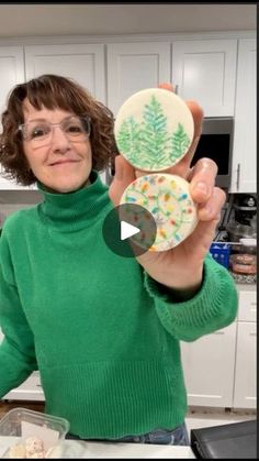 a woman holding up two decorated cookies in her hands and smiling at the camera while standing in front of a kitchen counter