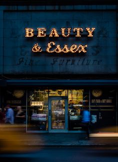 a store front with the word beauty and care lit up at night time, as pedestrians pass by