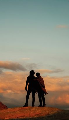 two people standing on top of a hill under a blue sky with clouds in the background