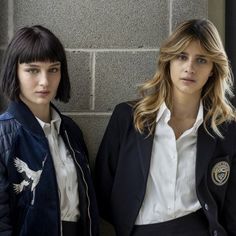 two women standing next to each other in front of a brick wall and wearing jackets