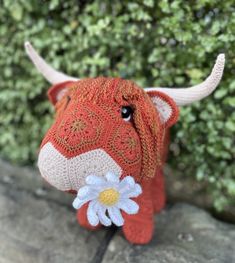 a knitted cow with a flower on its nose sitting in front of some bushes