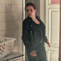 a woman standing in a bathroom next to a white sink and tiled walls, wearing a black leather jacket