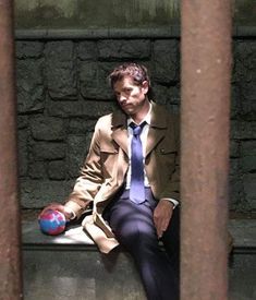 a man in a trench coat and tie sitting on a stone bench with a ball