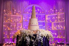 a wedding cake with white flowers on top in front of a purple lit display case