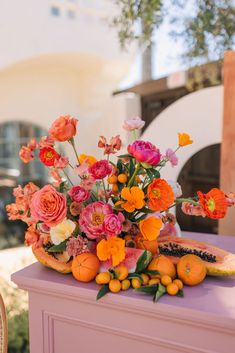 an arrangement of flowers and fruit on top of a pink table with chairs in the background