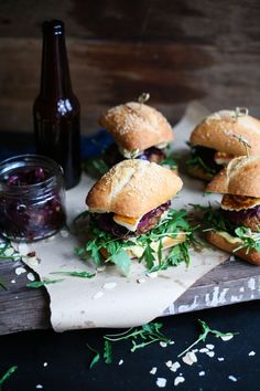 four sandwiches with meat and veggies sit on a cutting board next to a bottle of beer