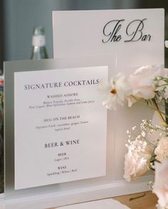 there is a sign that says signature cocktails on the table with flowers and wine bottles in the background