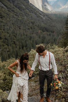 a man and woman are holding hands while walking through the woods with mountains in the background