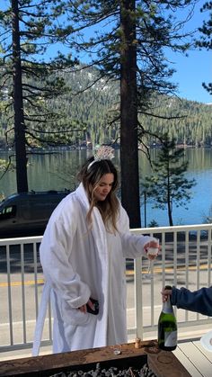 a woman in a robe pouring wine on a table with a lake and mountains in the background