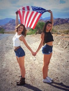 two young women holding hands while standing on the side of a road with an american flag