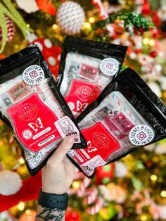 someone holding up two packets of christmas candy in front of a tree with ornaments on it