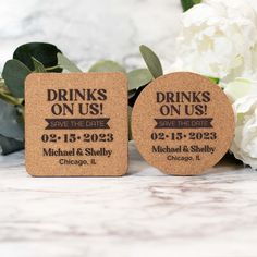 two cork coasters with drinks on us printed on them and flowers in the background