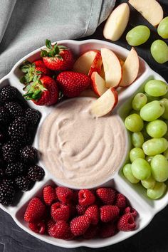 a white bowl filled with fruit and dip surrounded by grapes, apples, strawberries