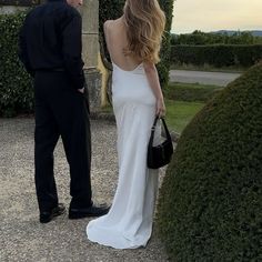 a man standing next to a woman in a white dress and holding a black purse