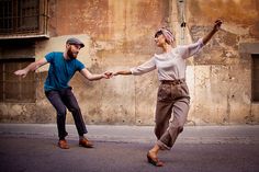 two people holding hands and dancing on the street in front of an old building with peeling paint