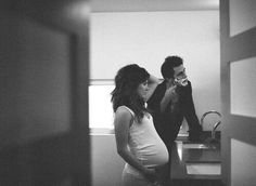 a pregnant woman standing next to a man in a kitchen sink while he brushes his teeth