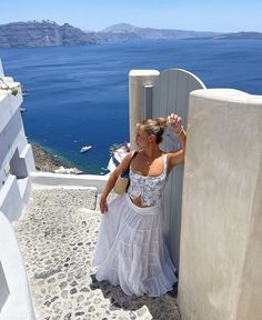 a woman in a white dress leaning against a wall with the ocean in the background