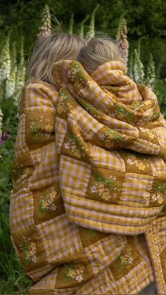 two children wrapped in blankets are sitting on a bench near tall grass and flowers, one is wearing a yellow plaid blanket