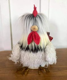 a stuffed chicken with long hair and a red bow on its head sitting on a wooden table