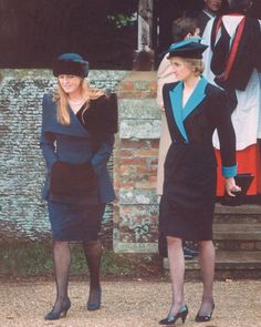 two women dressed in black and blue standing next to each other near a building with people looking on