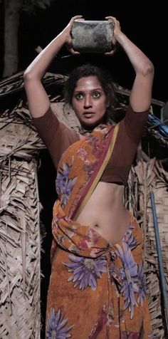 a woman in an orange sari is holding a pot over her head and looking at the camera