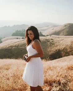 a woman in a white dress holding a camera on top of a grass covered hill