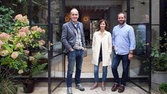 three people standing in front of an open glass door with plants and flowers behind them