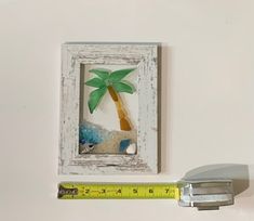 a white frame with a palm tree in it next to a measuring tape and a bottle opener