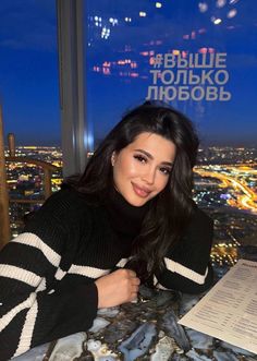 a woman sitting at a table in front of a window with the city lights behind her