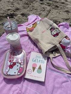 a hello kitty bag, book and drink on a towel at the beach with sand in the background