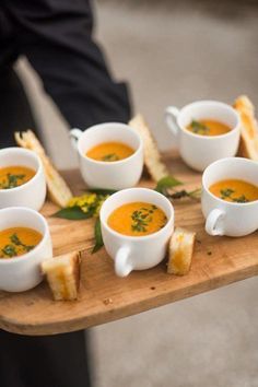 several cups of soup are on a wooden tray with bread slices and herbs in them