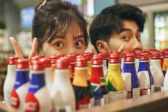 two people behind a row of bottles with one pointing at the camera and the other holding their fingers up