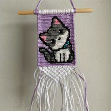 a purple and white wall hanging with a cat on it's side, next to a wooden stick